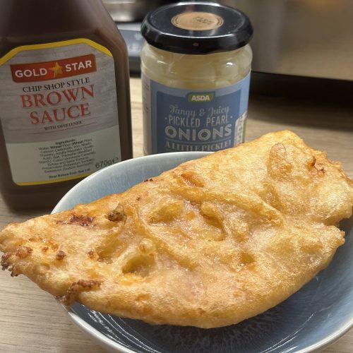 Cooked chip shop style pizza crunch on a plate with chip shop brown sauce and pickled onions on the side.