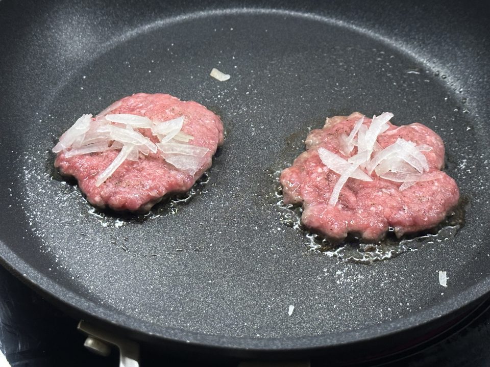Burger patties topped with sliced onion cooking in a frying pan.