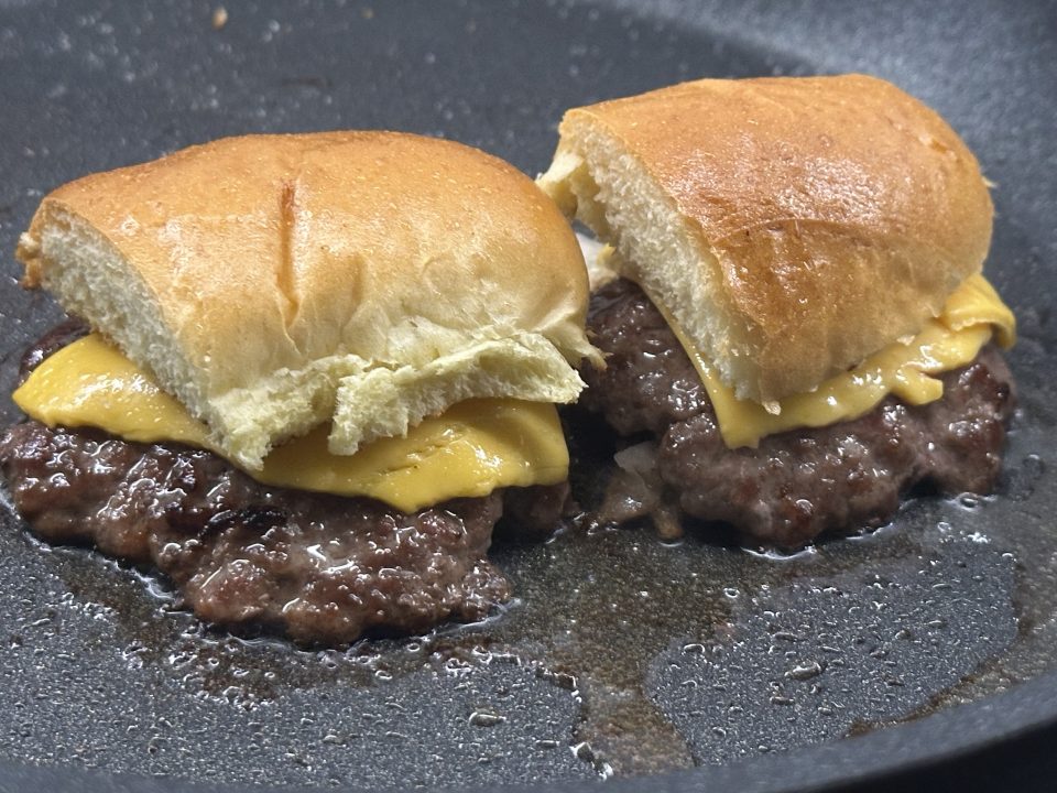 Cheeseburger sliders topped with top bun halves cooking in a frying pan.